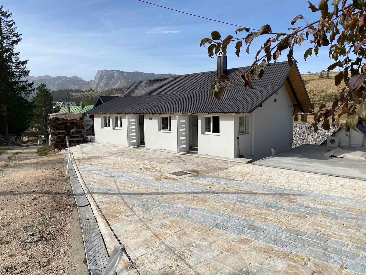 A modern cottage is situated on a flat stone patio, with mountains framed in the background. The building features a simple façade with two doors, and a gently sloped roof. Green trees are visible nearby, adding a natural touch to the surroundings.