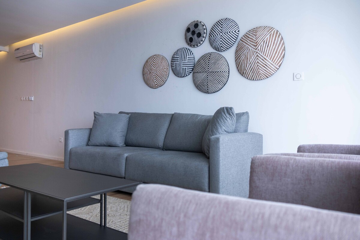 A light-filled living area features a gray sofa with assorted cushions, positioned under a decorative arrangement of patterned wall art. A dark coffee table is present, complementing the neutral tones of the space, which promotes a relaxed environment.