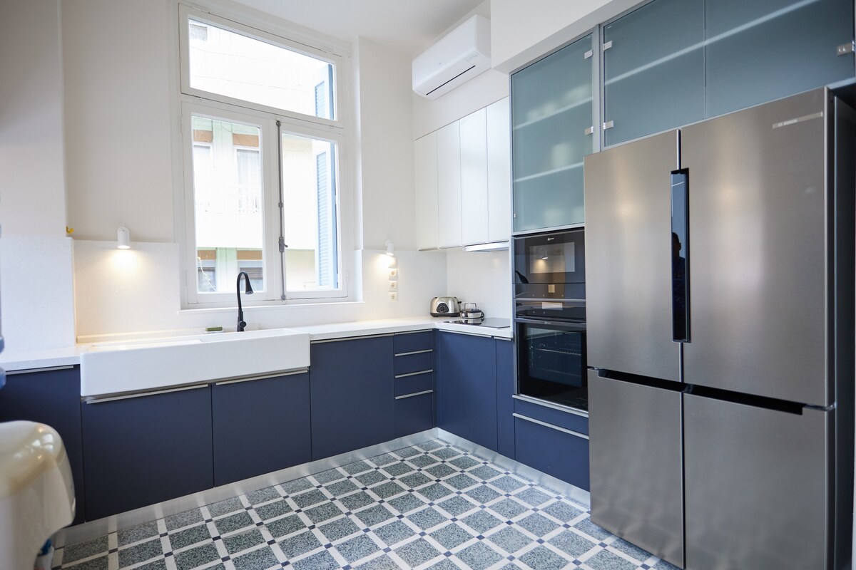 A modern kitchen features sleek cabinetry in two-tone blue and white finishes. A spacious sink and high-end appliances, including a stainless steel refrigerator and oven, are integrated into the design. Large windows allow natural light to brighten the room, showcasing a patterned tile floor.