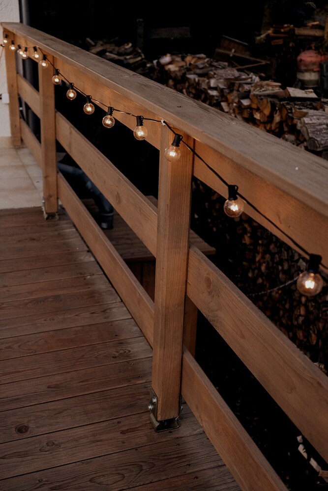 A wooden railing outlines a deck area, adorned with evenly spaced string lights that emit a warm glow. The surface of the deck is made of wooden planks, and the background reveals a stacked woodpile, enhancing the rustic ambiance.