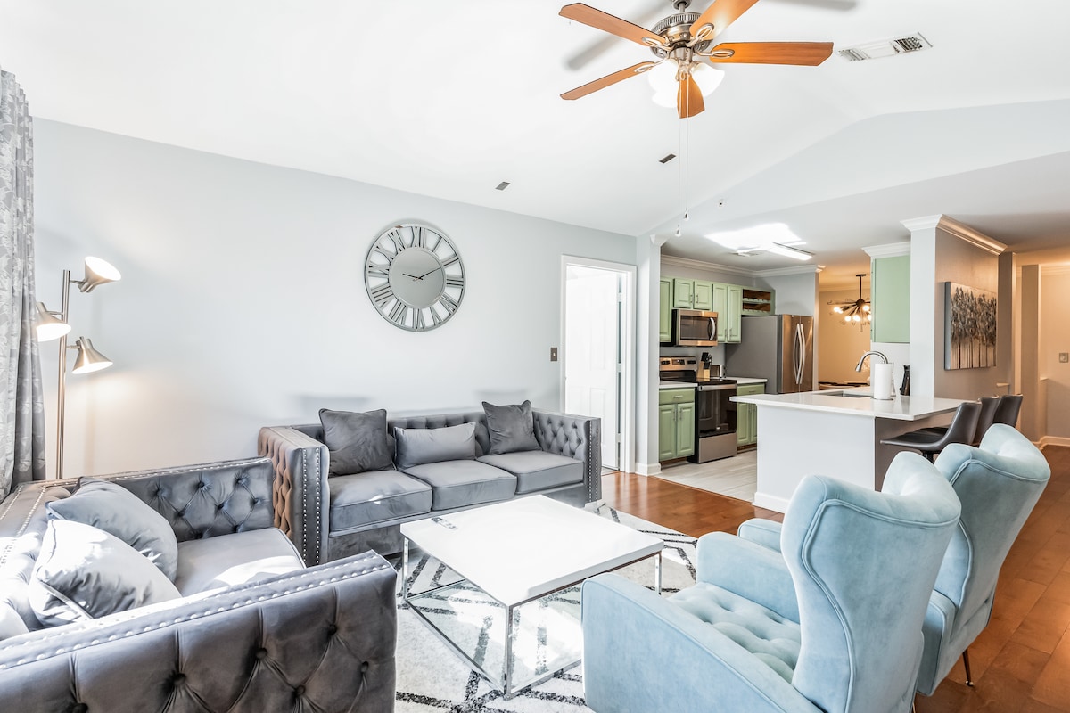 A spacious living area features modern furniture in shades of gray and blue. A large clock adorns the wall, while a glass coffee table serves as a central point. The kitchen is visible in the background, equipped with stainless steel appliances and green cabinetry.