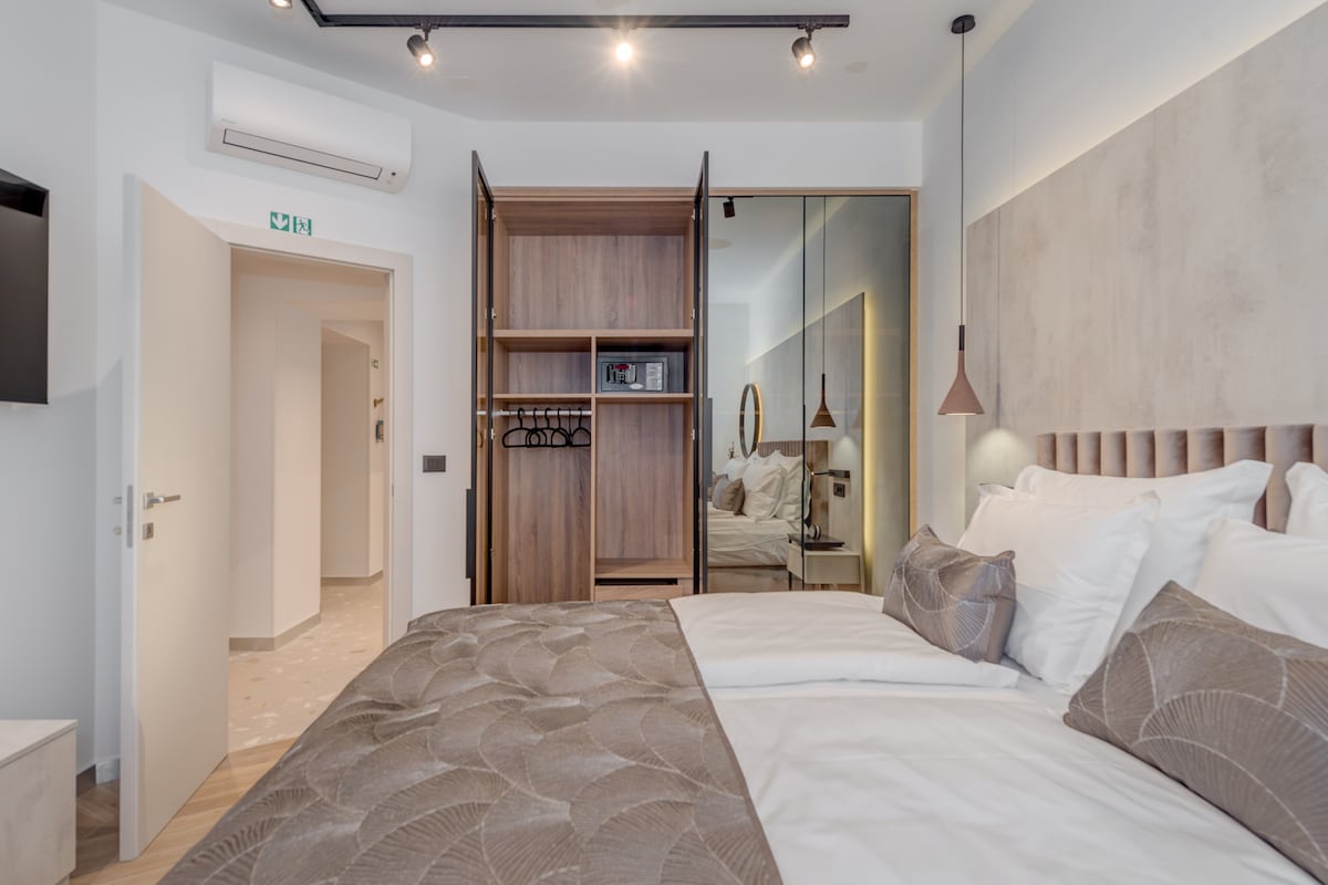 A well-appointed bedroom features a spacious bed with plush bedding and soft pillows. A mirrored wardrobe and stylish light fixture add elegance, while a subtle color palette creates a restful ambiance. An air conditioning unit and small wall-mounted television are present for comfort and convenience.