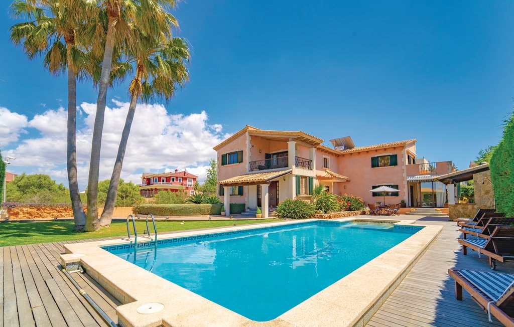 A spacious villa is shown, featuring a large swimming pool surrounded by a sun deck. Palm trees are visible alongside the pool area, and a shaded patio with outdoor seating is present. The two-story structure is painted in warm colors, set against a clear blue sky.
