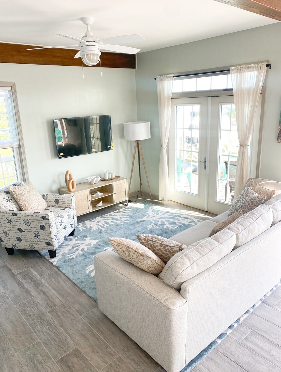 A living area features a light-colored sofa adorned with decorative pillows, paired with a patterned armchair. A flat-screen TV is mounted on the wall, while natural light fills the space through glass doors leading to the outdoor area. A neutral-toned area rug adds warmth.