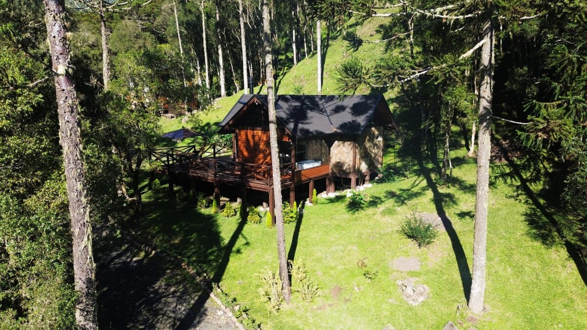 A charming chalet is situated amidst trees, with a wooden deck extending toward a grassy area. Surrounding nature creates a serene environment, while the structure features a combination of wood and stone elements. Natural light highlights the rustic design.