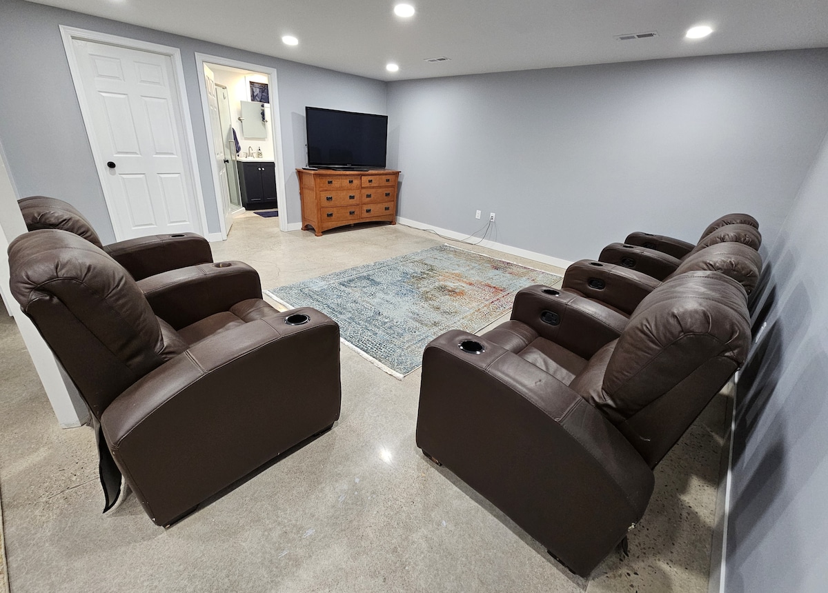 A cozy entertainment area features six reclining chairs arranged around a central rug. A wooden media console holds a television, and light gray walls create a calm setting. A doorway leads to another room, enhancing the spacious feel.