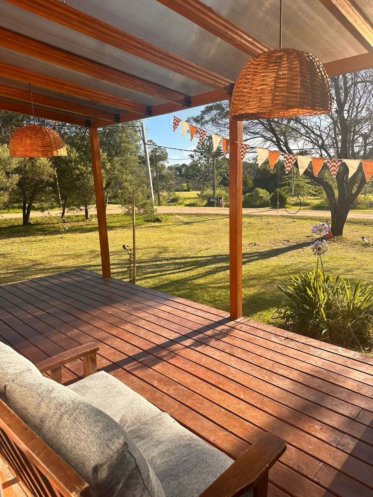 A wooden deck area is featured, highlighted by two woven pendant lights. A cozy seating arrangement with cushions provides a welcoming spot to enjoy the outdoor view. The surrounding greenery includes trees and a grassy lawn, enhancing the natural setting.