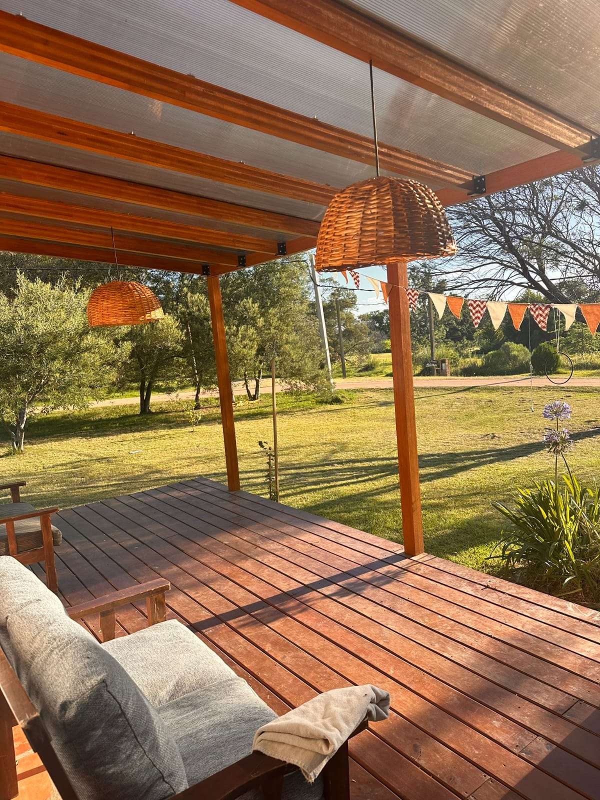 A spacious covered patio features wooden beams and a polished floor. Two woven hanging lights provide gentle illumination. A comfortable seating area is set on one side, complementing the serene garden view with greenery and decorative bunting across the space.