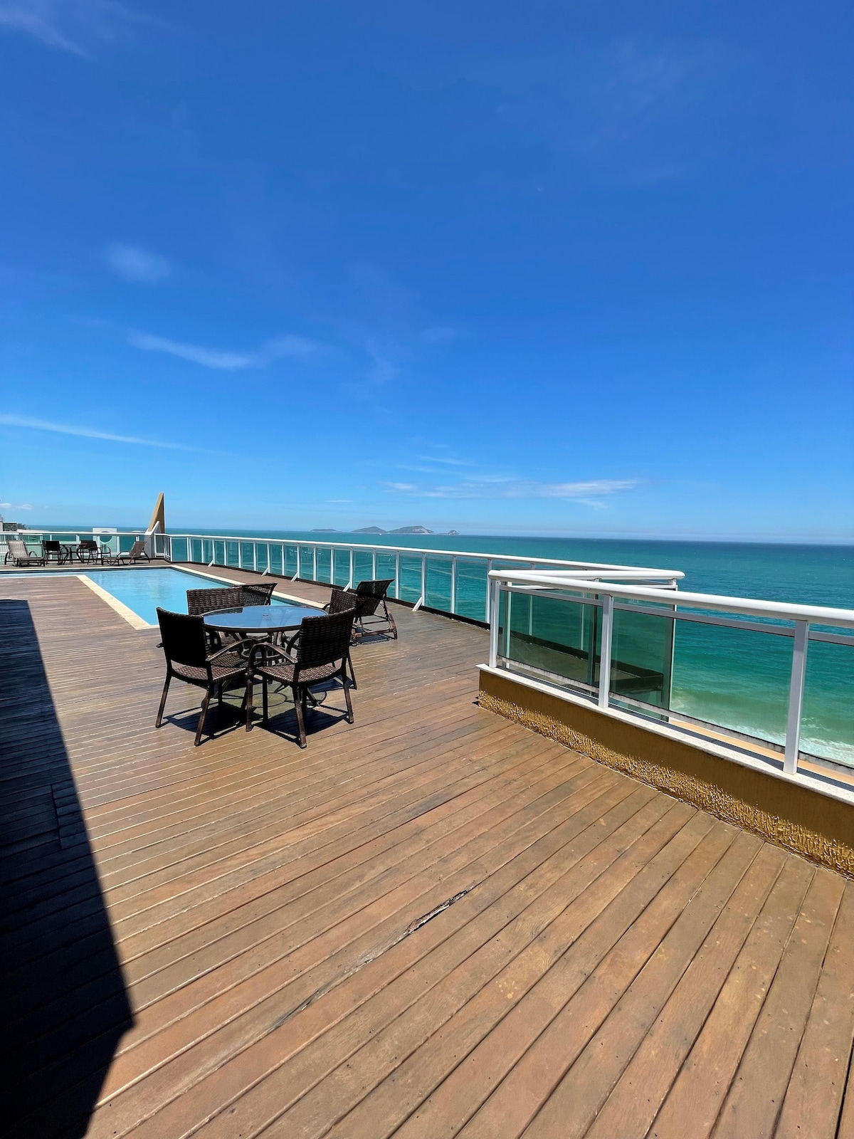 A sunlit outdoor deck features a polished wooden surface and a pool along the edge, offering panoramic ocean views. A dining set with four chairs provides a space for relaxation, while the clear sky complements the serene coastal setting.