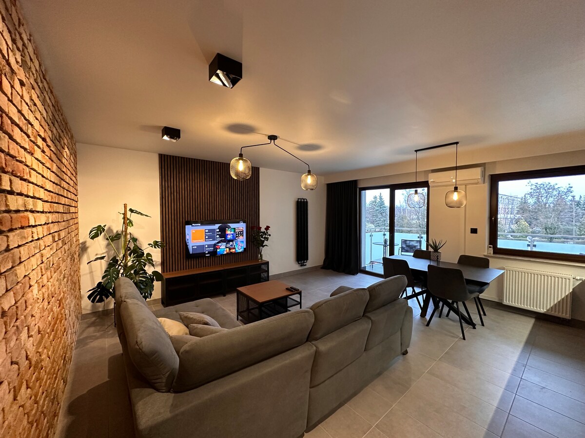 A modern living space is presented with a light grey sofa facing a wall-mounted television. Natural light fills the room through large windows, showcasing views of greenery. A wooden coffee table and dining area with chairs are also visible, contributing to a relaxed ambiance.