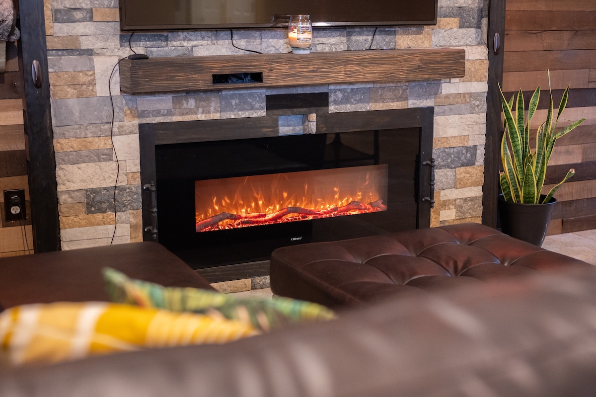 A modern electric fireplace is situated against a textured stone wall, providing a warm focal point in the living area. A stylish brown ottoman and a potted plant are visible in the foreground, enhancing the cozy ambiance of the space.