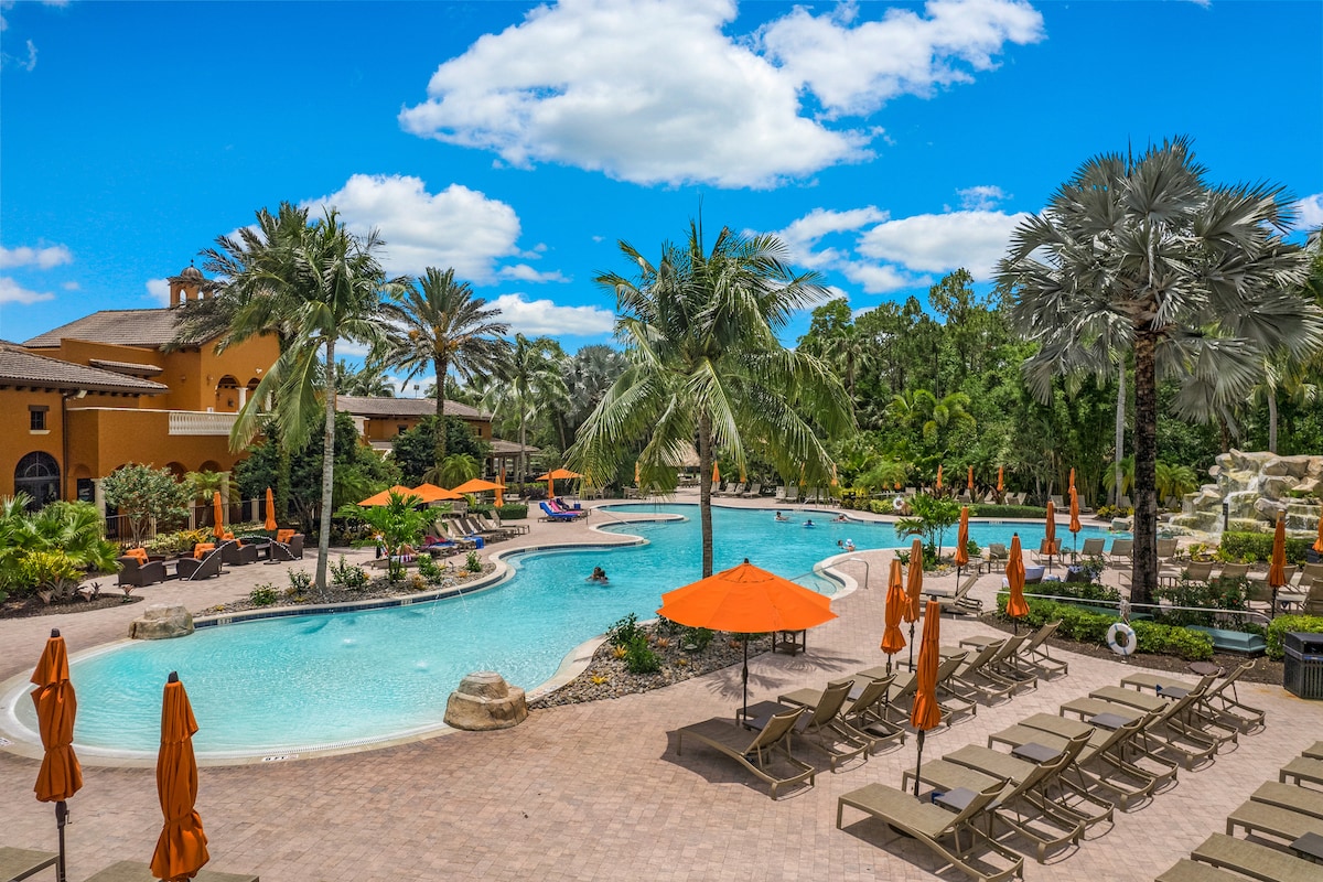 A resort-style pool area is surrounded by lush greenery and palm trees. Lounge chairs are arranged along the poolside, complemented by orange umbrellas providing shade. A waterfall feature and gentle slopes enhance the natural landscape, creating a serene setting for relaxation.