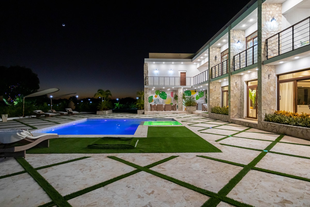 The villa is set against a twilight sky, showcasing a spacious outdoor area with a shimmering pool. Surrounding the pool, a stone patio is lined with neatly arranged grass and elegant loungers. The second level features large glass doors leading to a balcony, enhancing the views of the landscape.