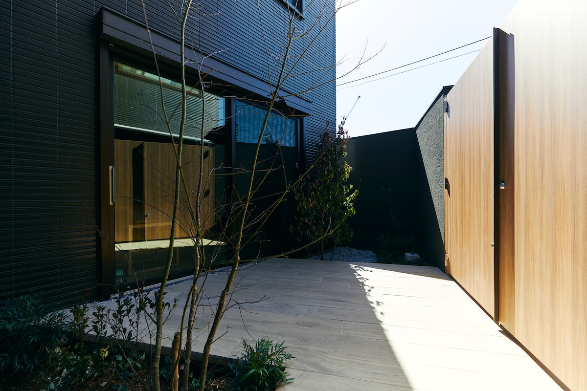 An entrance features a wooden gate opening to a small, private courtyard. Smooth wooden decking extends towards the home, flanked by minimal landscaping including greenery and stones, creating a serene outdoor space. Large windows provide glimpses of the interior's modern design.