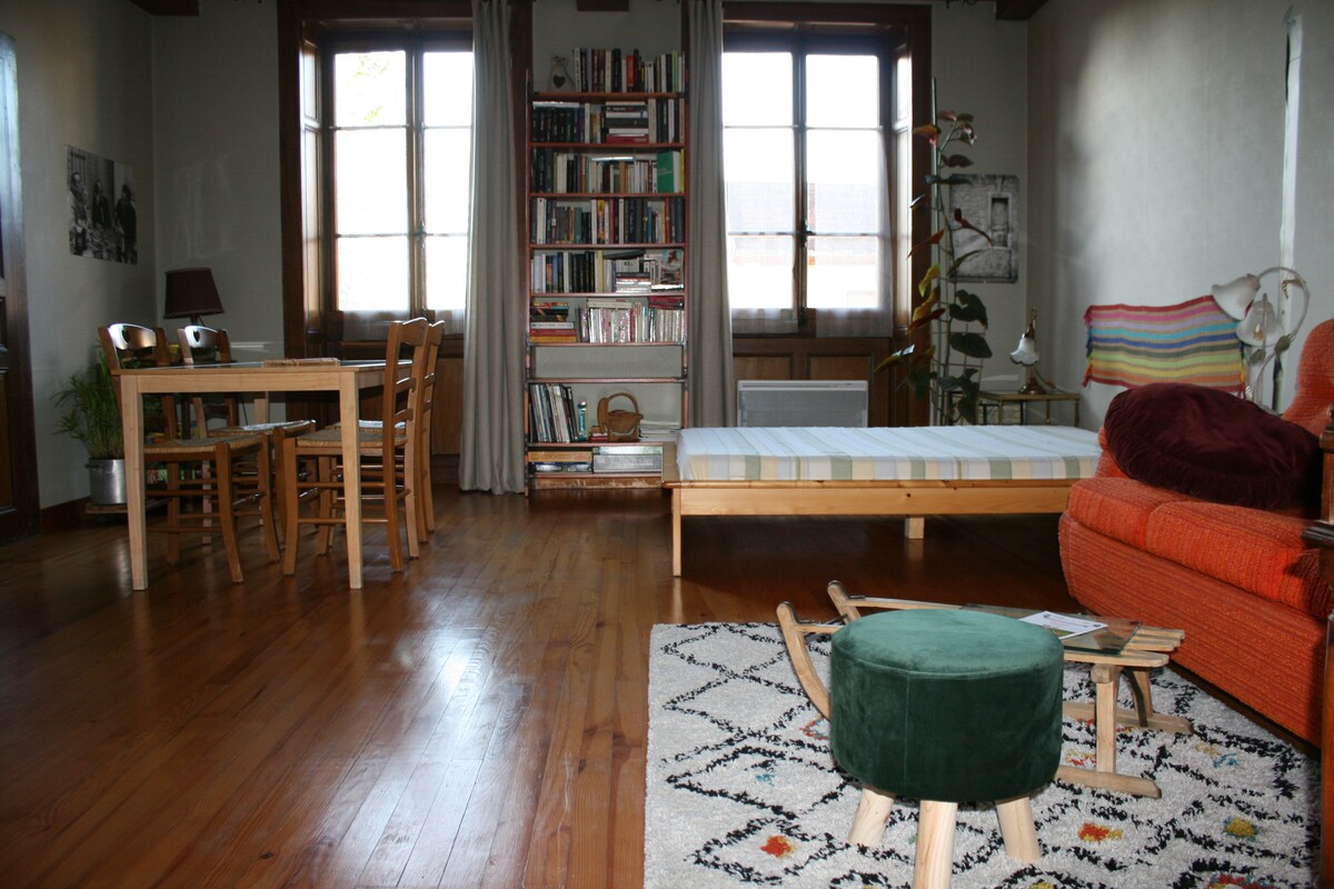 A spacious living area features a light wooden dining table with seating for four, a layered bookshelf filled with books, and a cozy orange couch. A striped bed is placed against the wall, with a traditional rug beneath a green pouf and wooden rocking chair.