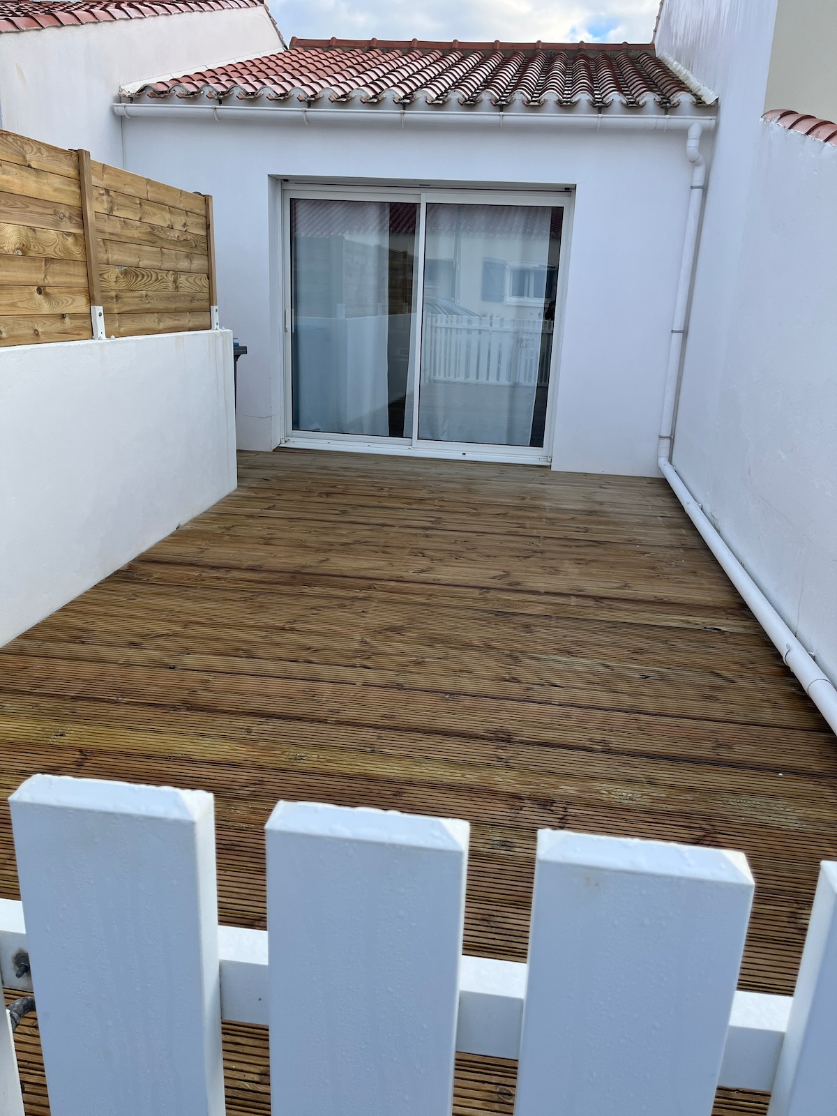 A wooden terrace features smooth planks, providing an outdoor seating area. A sliding glass door opens to the interior, allowing natural light to enter. Whitewashed walls create a clean, minimalistic look, and a fenced section adds privacy.
