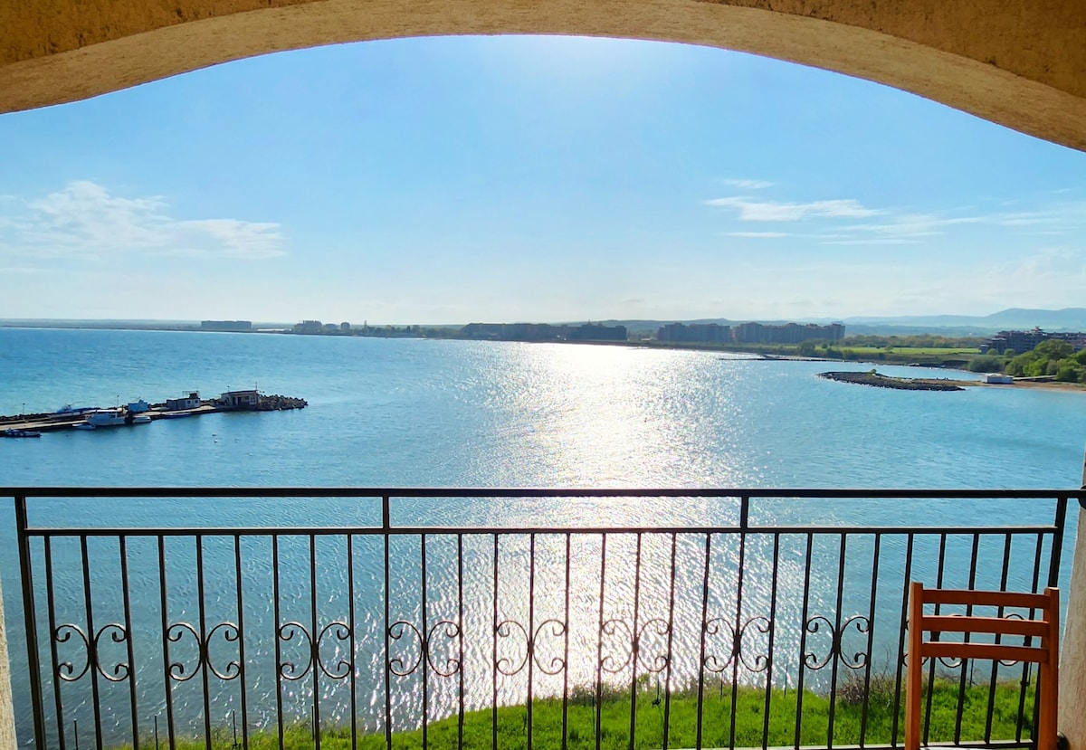 A panoramic view of the shimmering water is framed by an arched balcony. The light reflects off the surface, creating a tranquil atmosphere. A single wooden chair is positioned for enjoying the scenic vista, with distant green hills and a coastline visible.