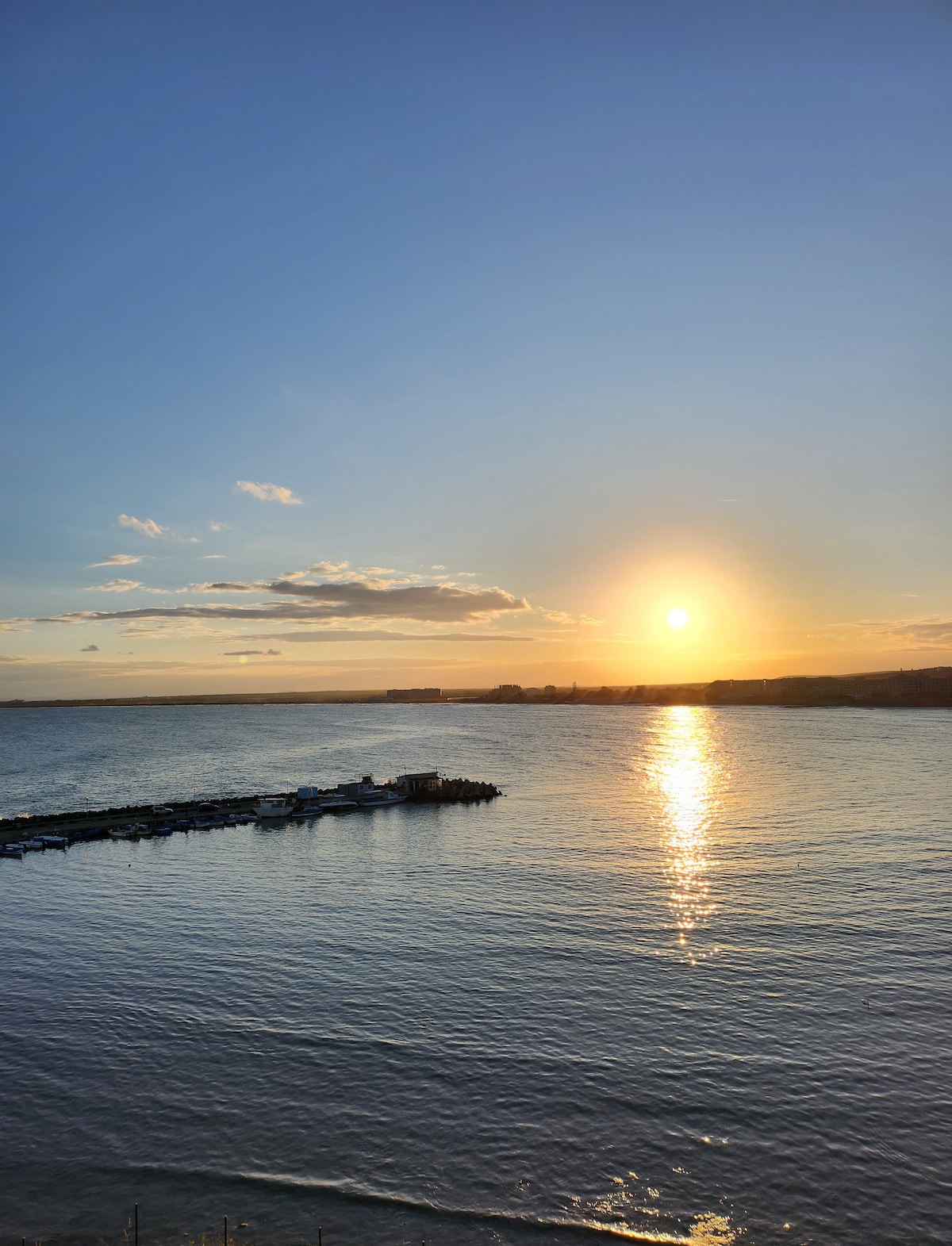A serene waterfront scene captures the sun setting over calm waters. The golden rays reflect on the surface, creating a warm glow. A dock extends into the water, providing a sense of connection to the landscape, while a gentle horizon line meets a clear blue sky.