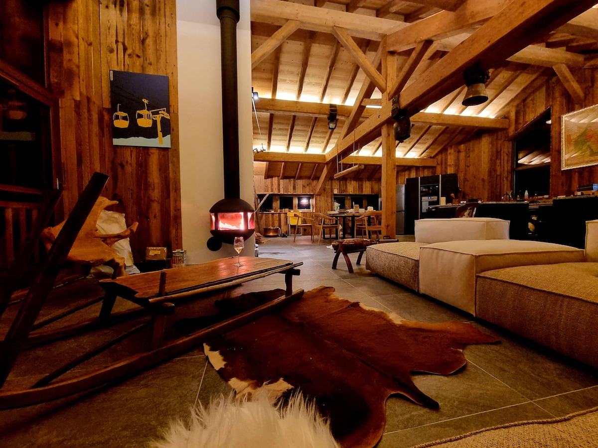 A warm and inviting living area features wooden beams and a cozy stove. A soft, light-colored seating arrangement is complemented by a rustic coffee table. A fur rug sits on stone flooring, while the open kitchen and dining space are visible in the background.