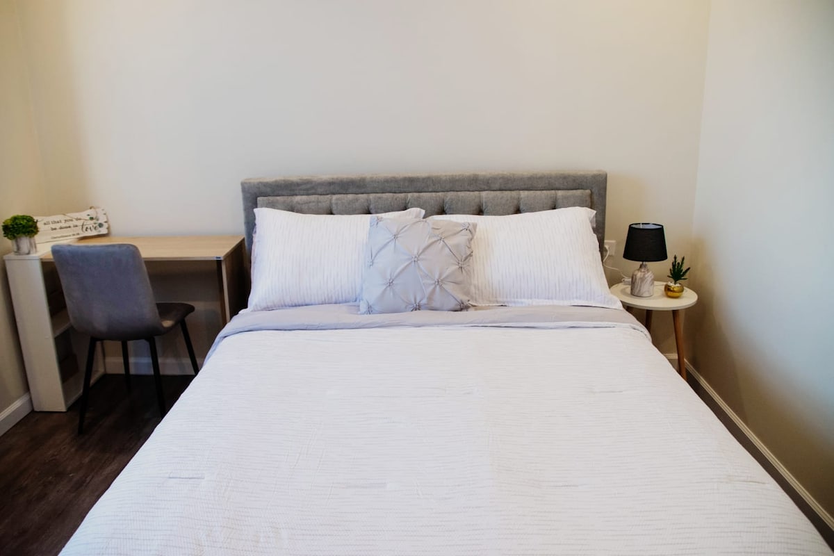 A spacious sleeping area features a neatly made bed with light-colored linens and decorative pillows. A small bedside table with a lamp and a modest plant is present. A desk and chair are situated to one side, providing a functional workspace.