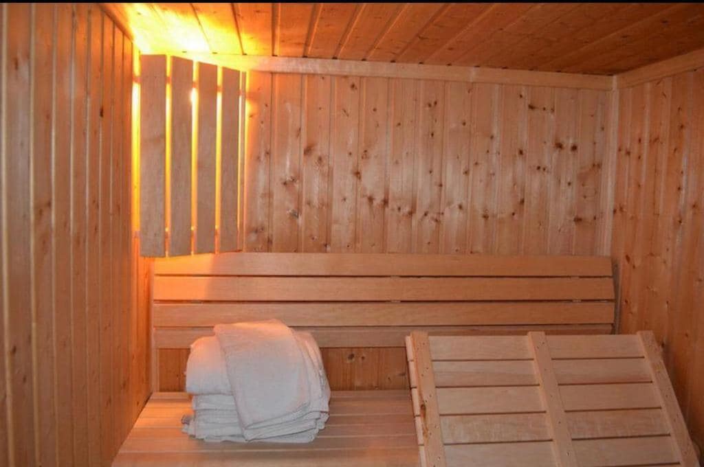 The interior of a sauna is presented with warm wooden paneling and benches. Soft, folded towels are neatly arranged, and gentle light filters in through a slatted opening, creating a soothing ambiance in the space.