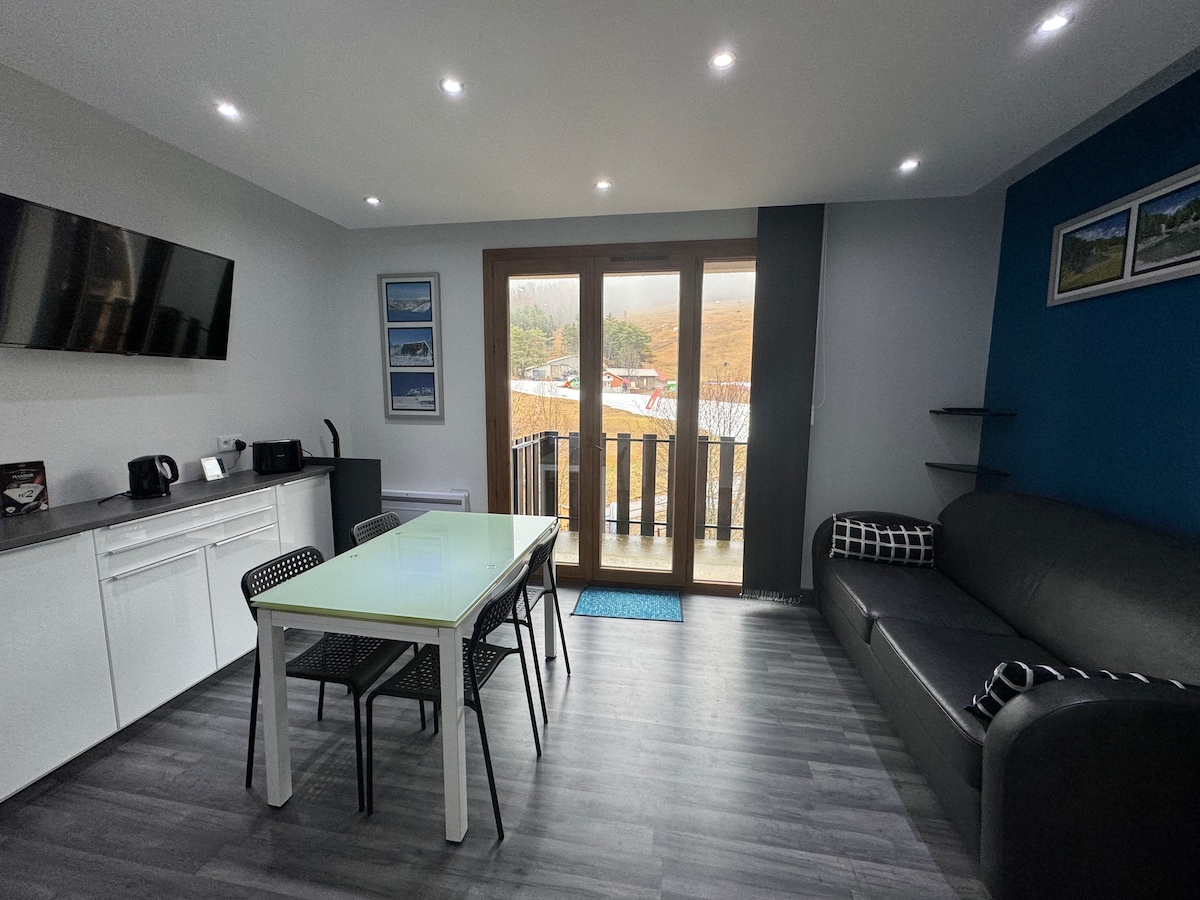 A sleek living area features a dining table accommodating four, complemented by modern black chairs. A comfortable sofa is positioned beside large glass doors that open to a balcony, allowing natural light to fill the space. A flat-screen TV is mounted on the wall.