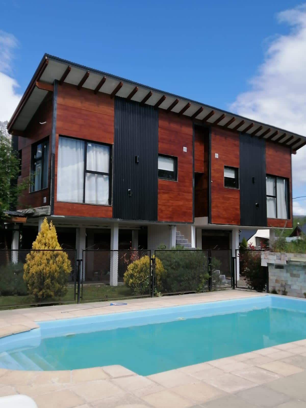 A modern two-story building showcases a combination of wooden and black paneling, set against a clear blue sky. A serene pool is visible in the foreground, bordered by a stone patio and surrounded by neatly landscaped greenery.