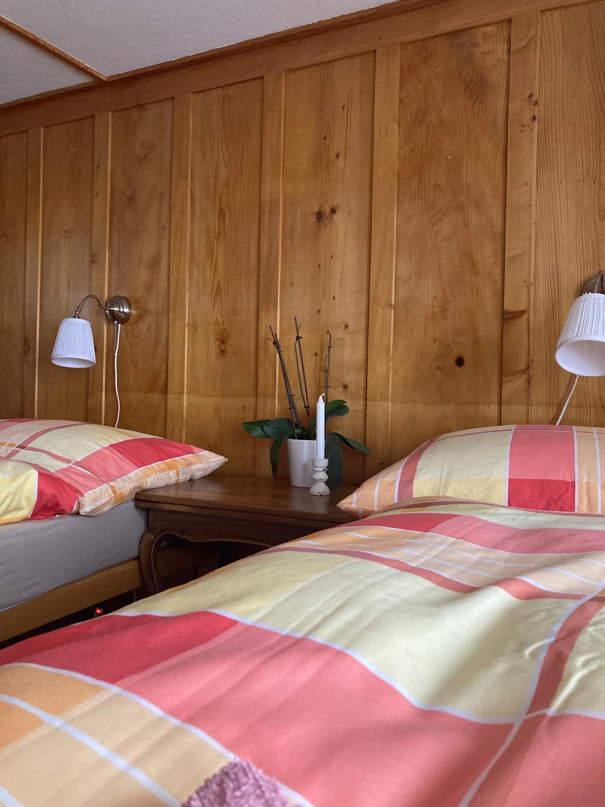 A cozy bedroom features two beds dressed in patterned quilts. Each bed is accompanied by a wall-mounted lamp and a small nightstand holding a candle and a plant. The warm wooden paneling enhances the room's inviting ambiance.