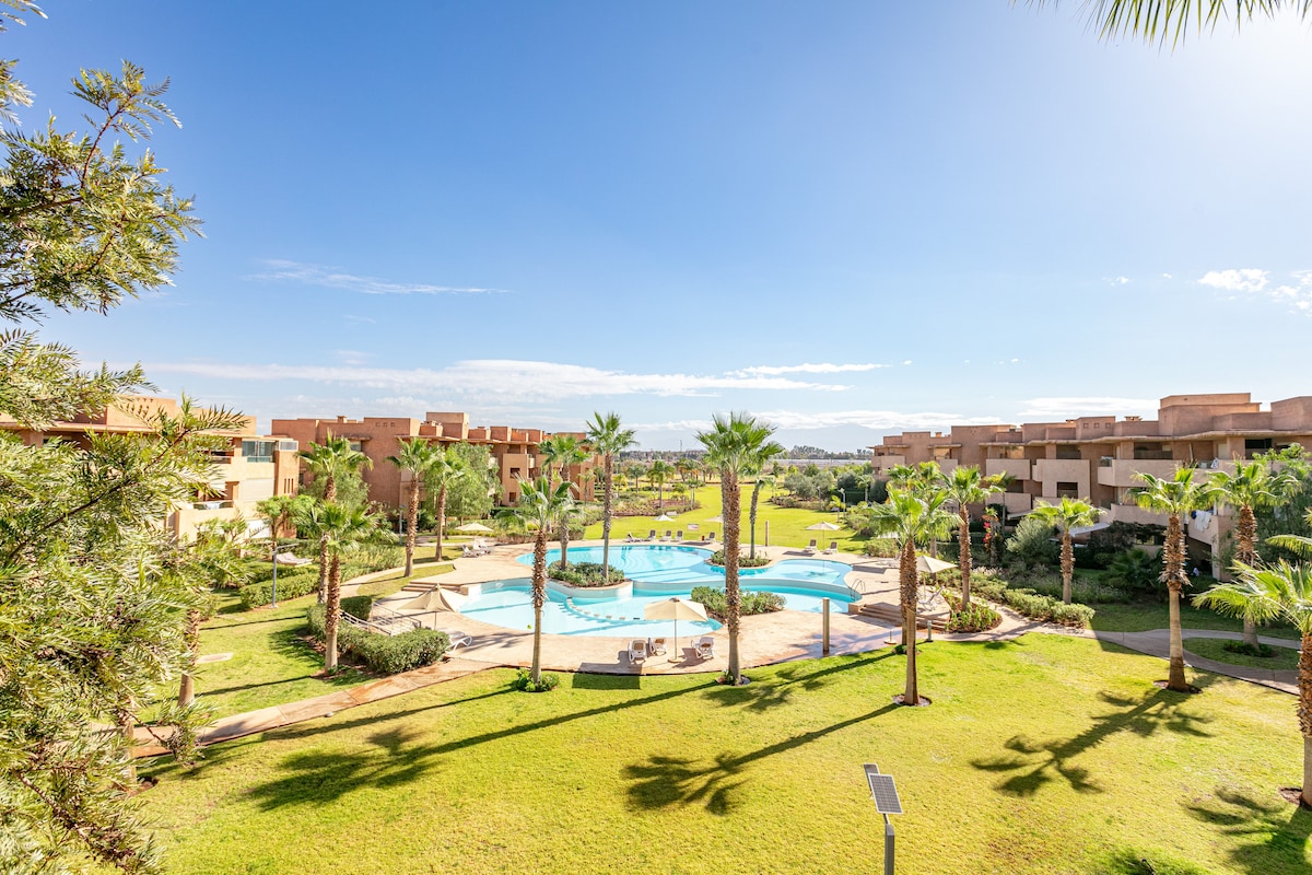 A serene view of the landscaped grounds showcases two swimming pools surrounded by palm trees and manicured lawns. The modern architecture of the neighboring buildings complements the lush greenery under a clear blue sky.