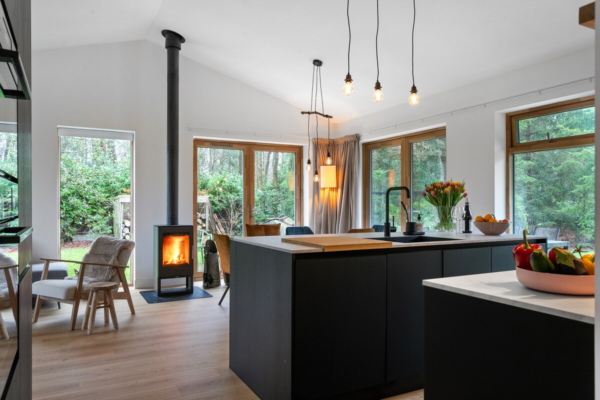 The kitchen area features a modern design with an island and wood-burning stove, creating a central gathering space. Bright natural light streams through large windows, illuminating fresh flowers and an array of fruits on the countertop. Comfortable seating arrangements are positioned nearby.