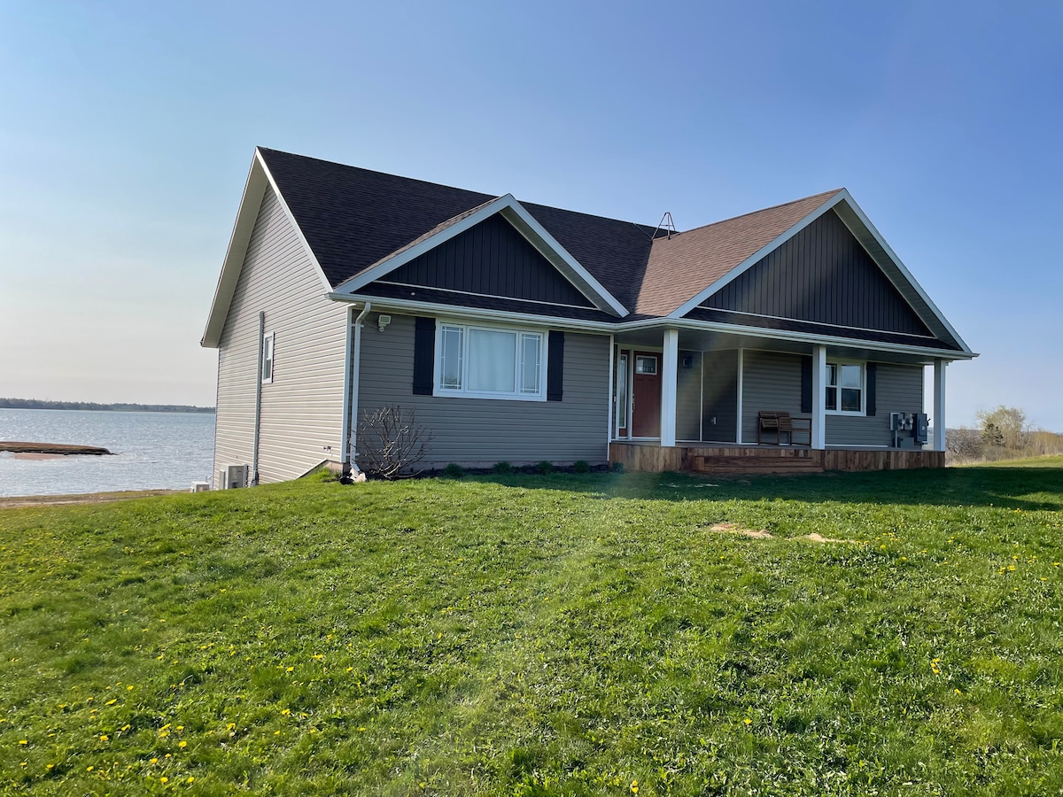 The house is set against a scenic waterfront backdrop, featuring a wide expanse of grass leading to the water's edge. Its exterior is composed of neutral siding, with a combination of large windows and a covered porch, inviting relaxation and enjoyment of the surroundings.