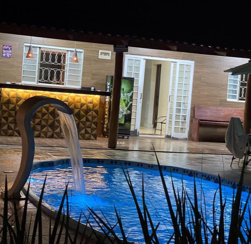 A serene backyard features a softly illuminated pool with a waterfall feature. A nearby outdoor bar area includes warm lighting, and the entrance to the house is framed by double glass doors. Potted plants enhance the tranquil setting, contributing to a calming atmosphere.