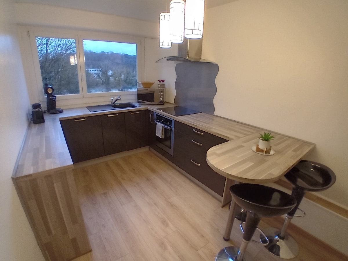 The kitchen features modern cabinetry with wood laminate surfaces, equipped with an electric stove, oven, and coffee maker. A small round table with two black bar stools is placed near the window, offering natural light and a view of the surroundings.