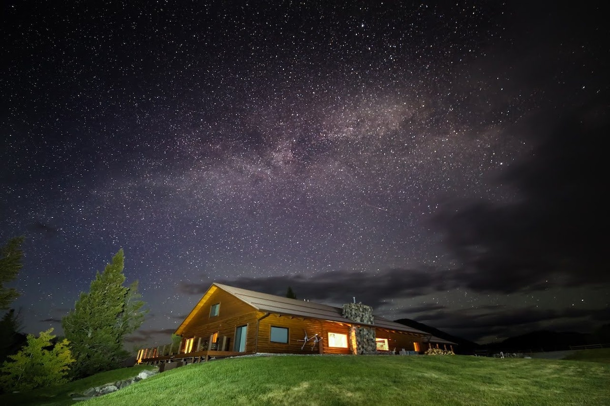 A spacious log home is depicted against a starlit sky, featuring warm light emanating from its windows. Lush green grass surrounds the building, while trees are present in the foreground, contributing to the serene natural setting.