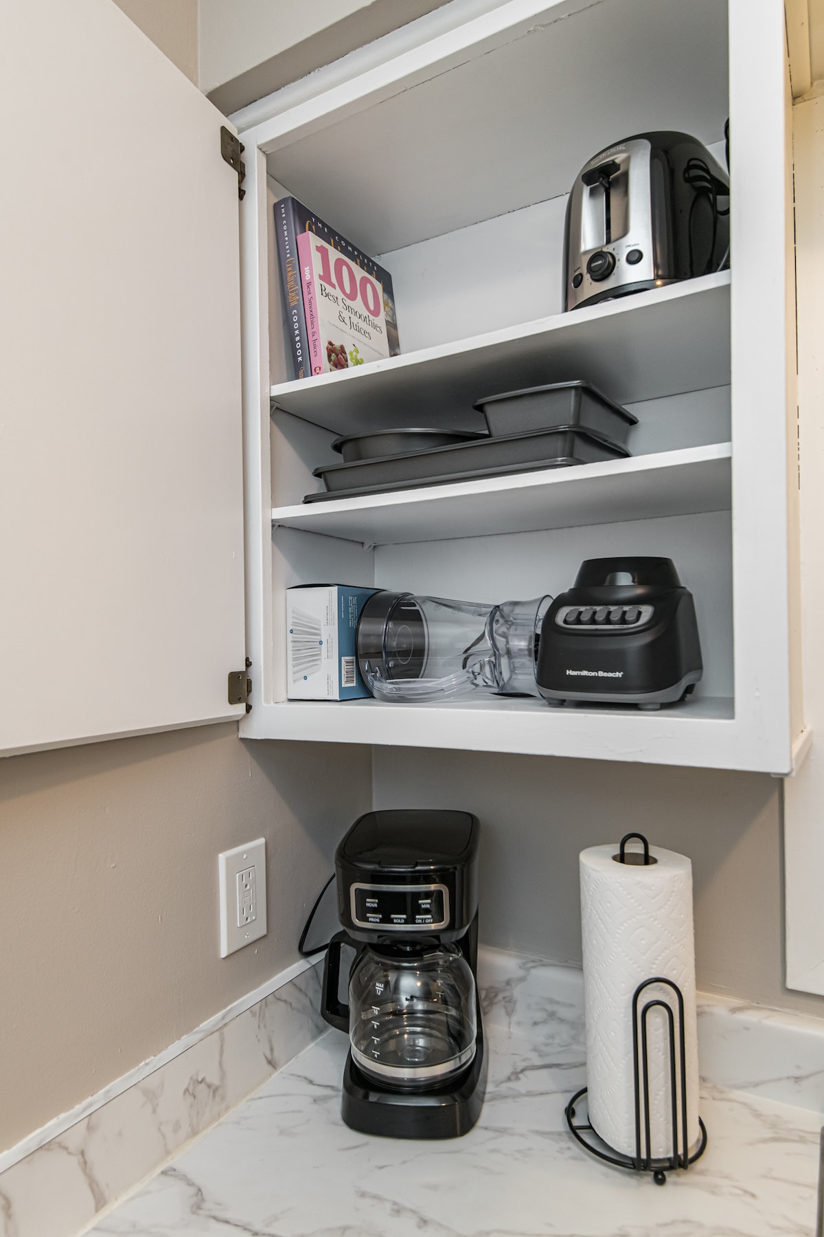 A well-organized cabinet displays various kitchen appliances, including a toaster, a blender, and a coffee maker. A cookbook titled '100' is visible on the shelf, along with a set of baking pans. Paper towels are neatly placed in a holder next to the coffee maker.