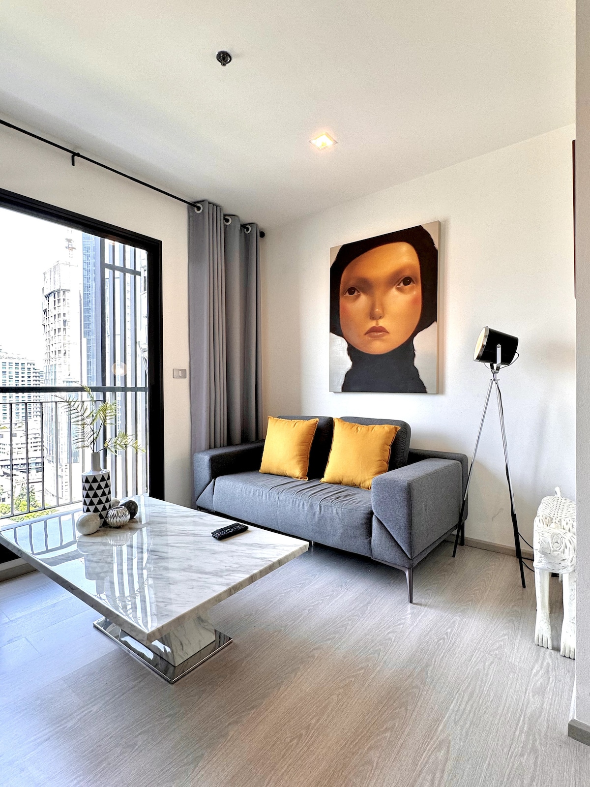 A modern living area features a gray sofa adorned with two yellow cushions, complemented by a marble coffee table. Natural light filters through a large window, revealing a glimpse of the cityscape. Grey curtains frame the window, adding a refined touch to the space.