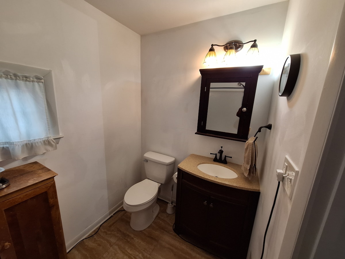 The private bathroom features a modern toilet and a sleek vanity with a circular sink. Above the vanity, a mirror is framed in dark wood, and warm lighting is provided by fixtures with three bulbs. A towel hangs conveniently from a hook, enhancing functionality.