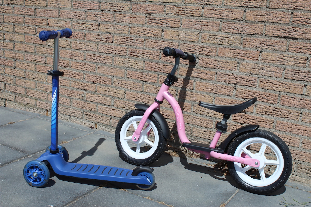 A blue kick scooter and a pink balance bike are positioned on a paved surface against a textured brick wall. The balance bike features two wheels and a comfortable seat, while the scooter has a sturdy platform and adjustable handlebar.
