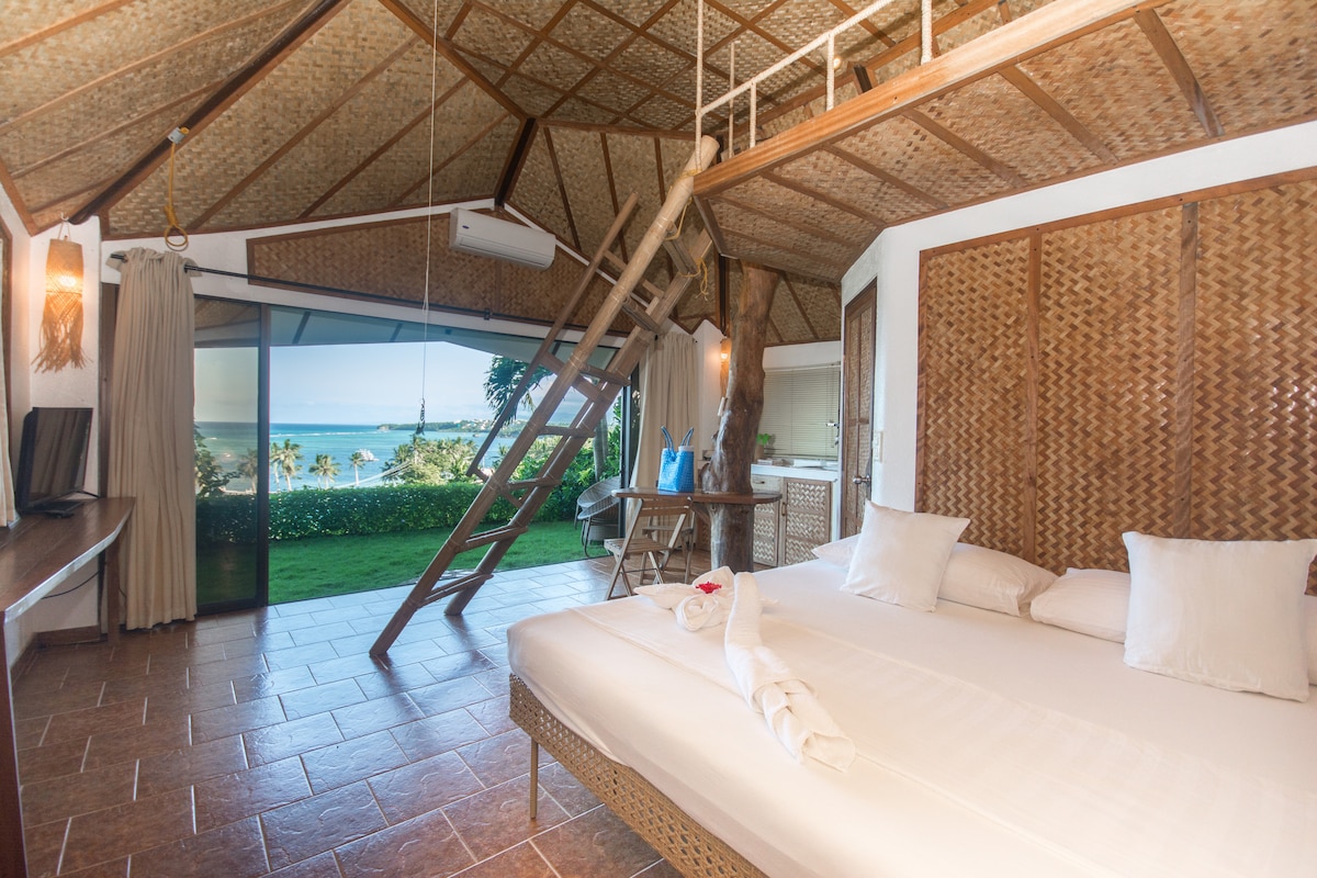 A cozy bungalow interior features a spacious king bed with soft linens and a loft bed above. Large sliding glass doors provide a panoramic view of the ocean, allowing natural light to brighten the room. Warm, textured walls enhance the tropical ambiance.