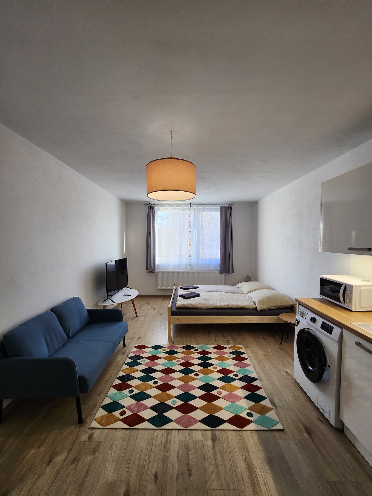 The living space features a cozy double bed positioned beside a large window with sheer curtains, allowing natural light to filter in. A blue sofa and a colorful patterned rug enhance the warm atmosphere. A washing machine and kitchenette are visible along one wall.