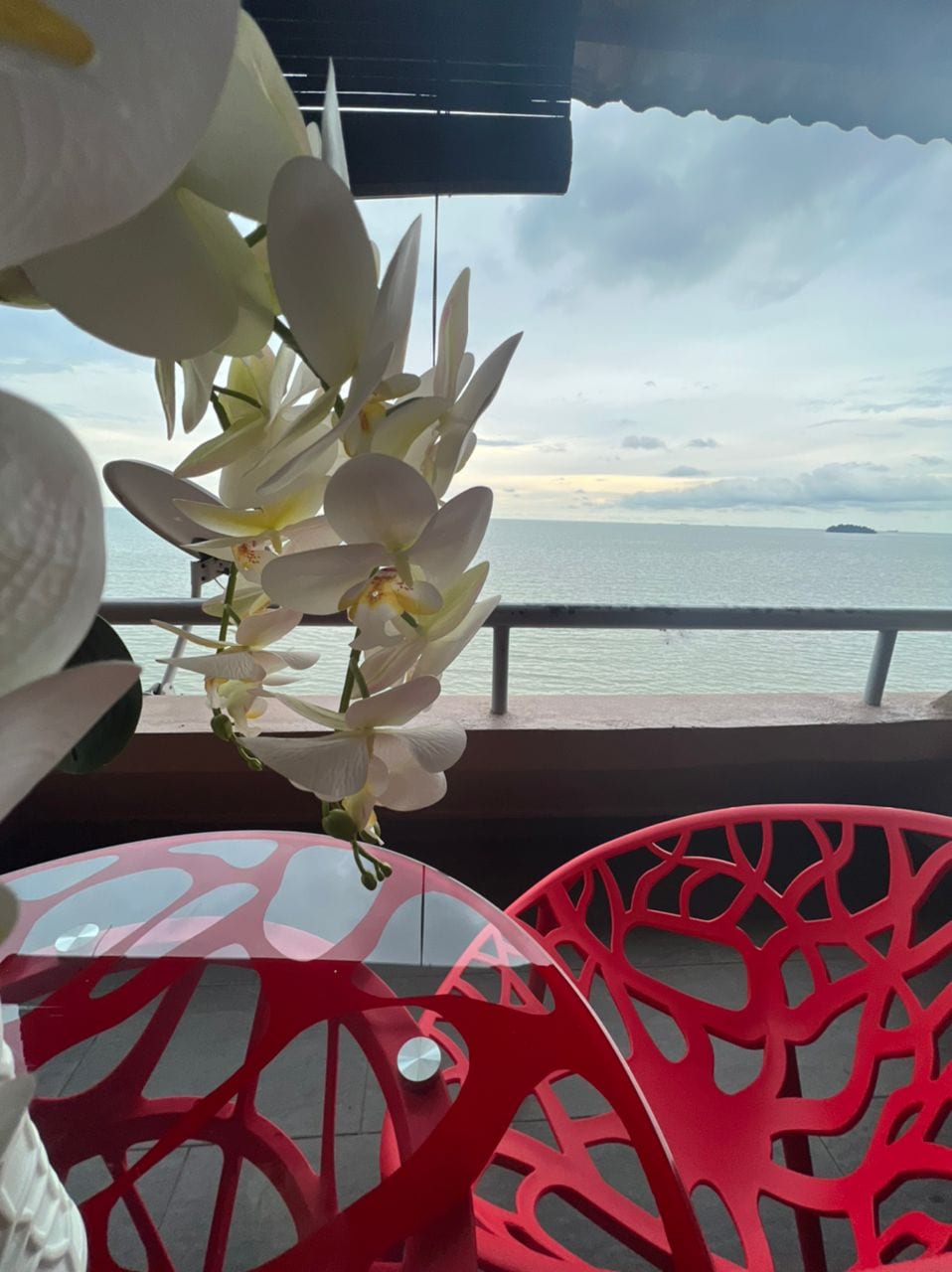 A dining area is displayed with a clear glass table and two vibrant red chairs featuring intricate, organic designs. White orchids are positioned in the foreground, while a calm sea and distant islands are visible through a balcony railing.