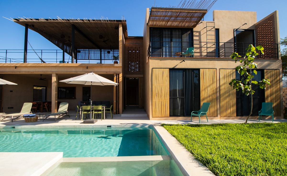 A modern villa is presented with a clear pool in the foreground. The structure features a blend of natural materials and large windows. A spacious balcony is visible on the upper floor, with outdoor seating and umbrellas providing shade in the garden area.