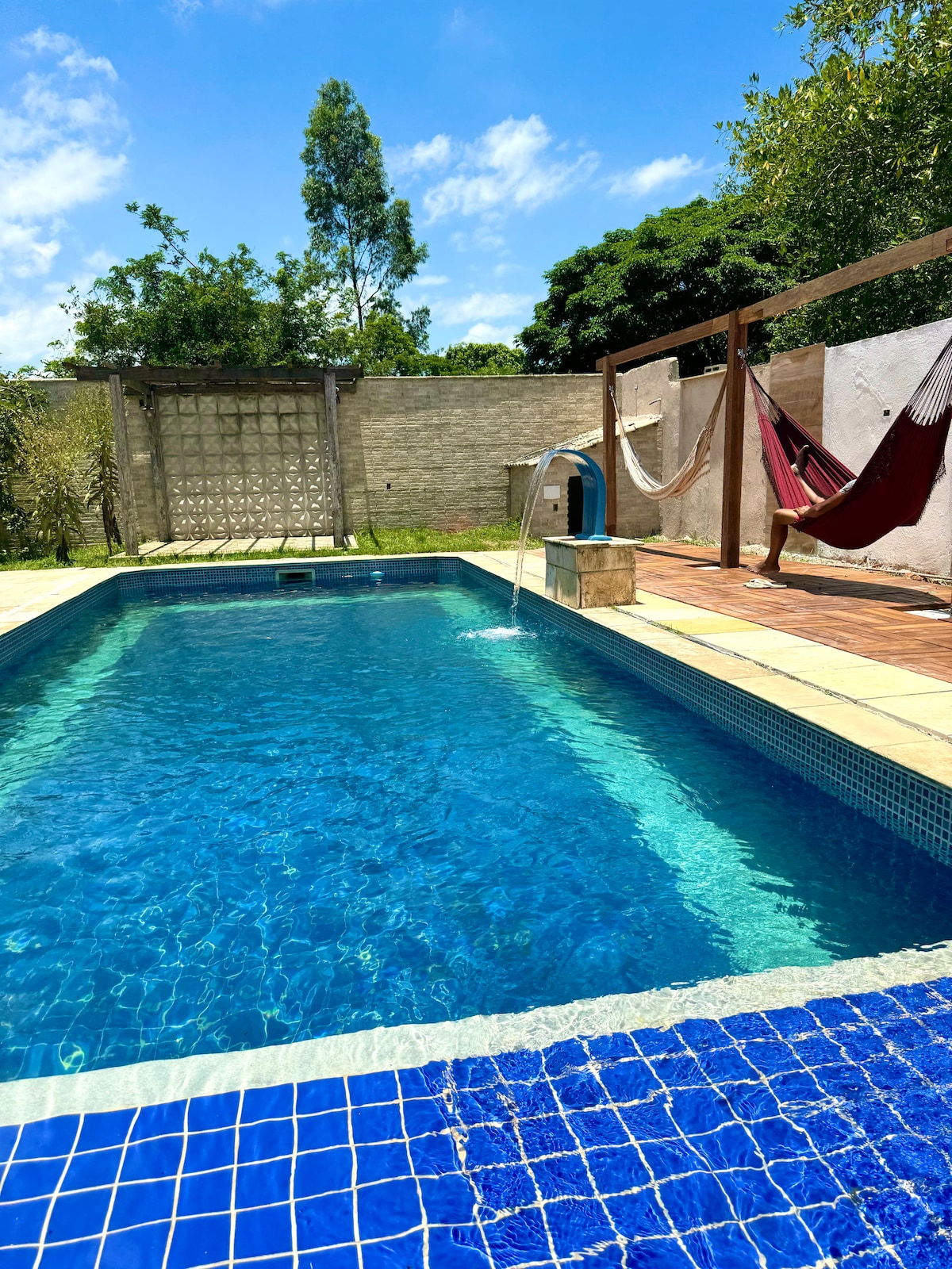 A clear swimming pool is surrounded by a tiled deck, featuring a gentle waterfall feature. Nearby, two hammocks are suspended from wooden beams, inviting relaxation. Lush greenery and a light blue sky with scattered clouds are visible in the background, enhancing the tranquil outdoor setting.