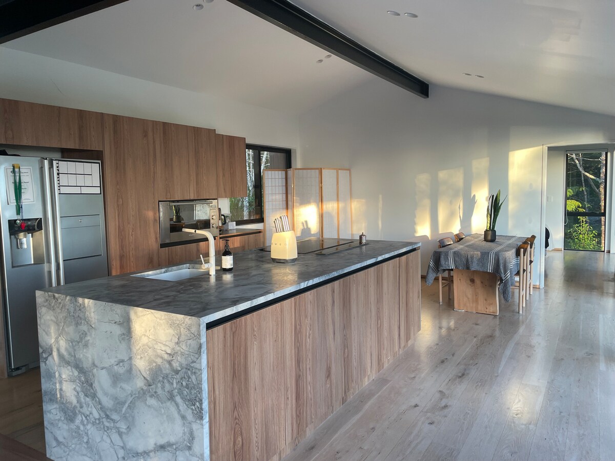 A spacious kitchen and dining area showcases a large marble-topped island with wooden cabinetry. Natural light filters in through large windows, illuminating a simple dining table set for six. A light-colored throw and decorative plants are placed on the side.