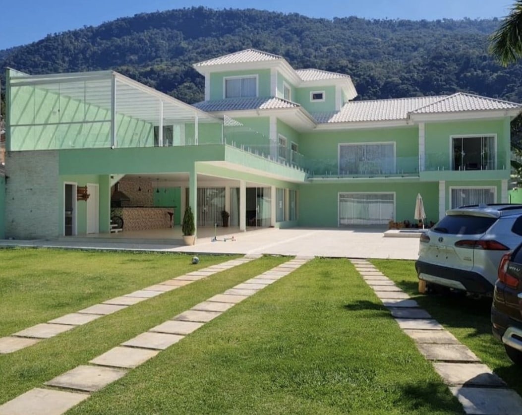 A spacious green house with multiple levels and large windows stands against a mountain backdrop. The exterior features a wide open patio area surrounded by manicured grass and a stone pathway leading to the entrance. Sunlight brightens the modern architecture.