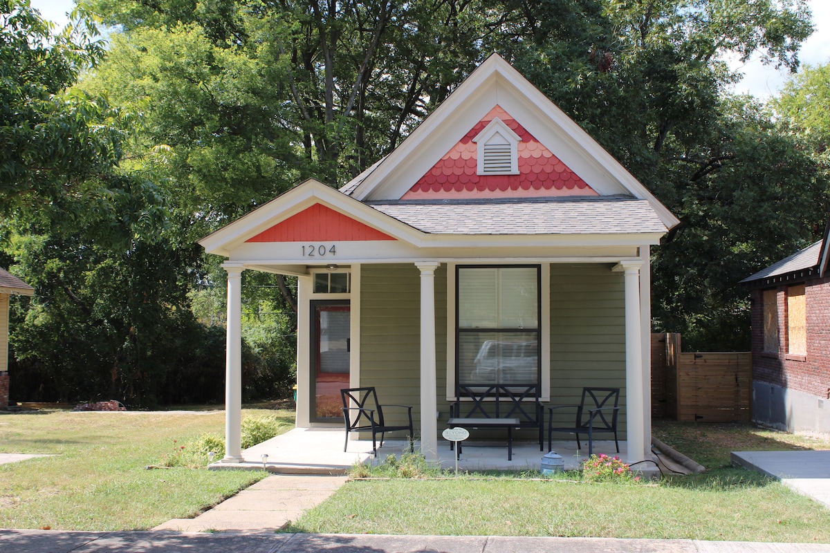 A charming exterior view of a single-story house features a front porch with two black chairs, welcoming guests. The house showcases a blend of green siding and red shingles, with a distinct wooden door and a large window allowing natural light.