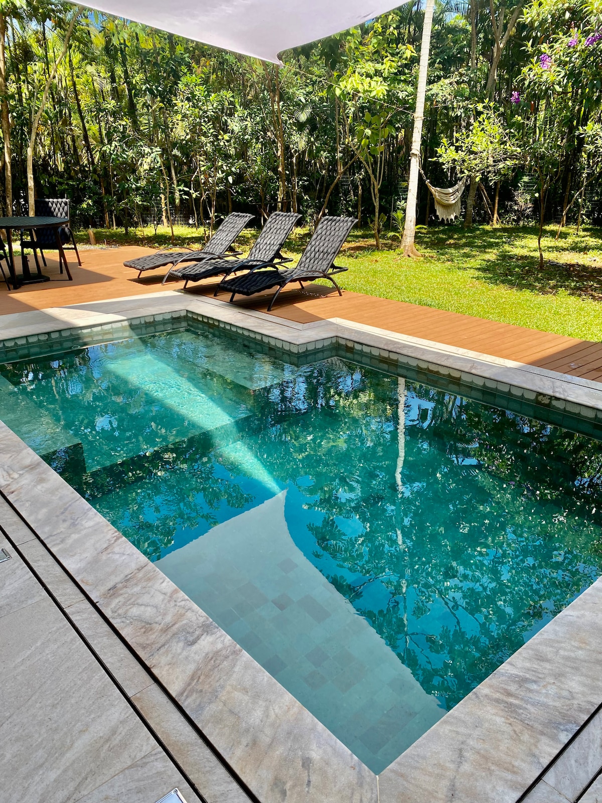 A private pool is set in a natural outdoor area, surrounded by lush greenery. Four lounge chairs are positioned alongside the pool on a wooden deck. A dining table is visible nearby, providing an inviting space for relaxation and enjoyment.