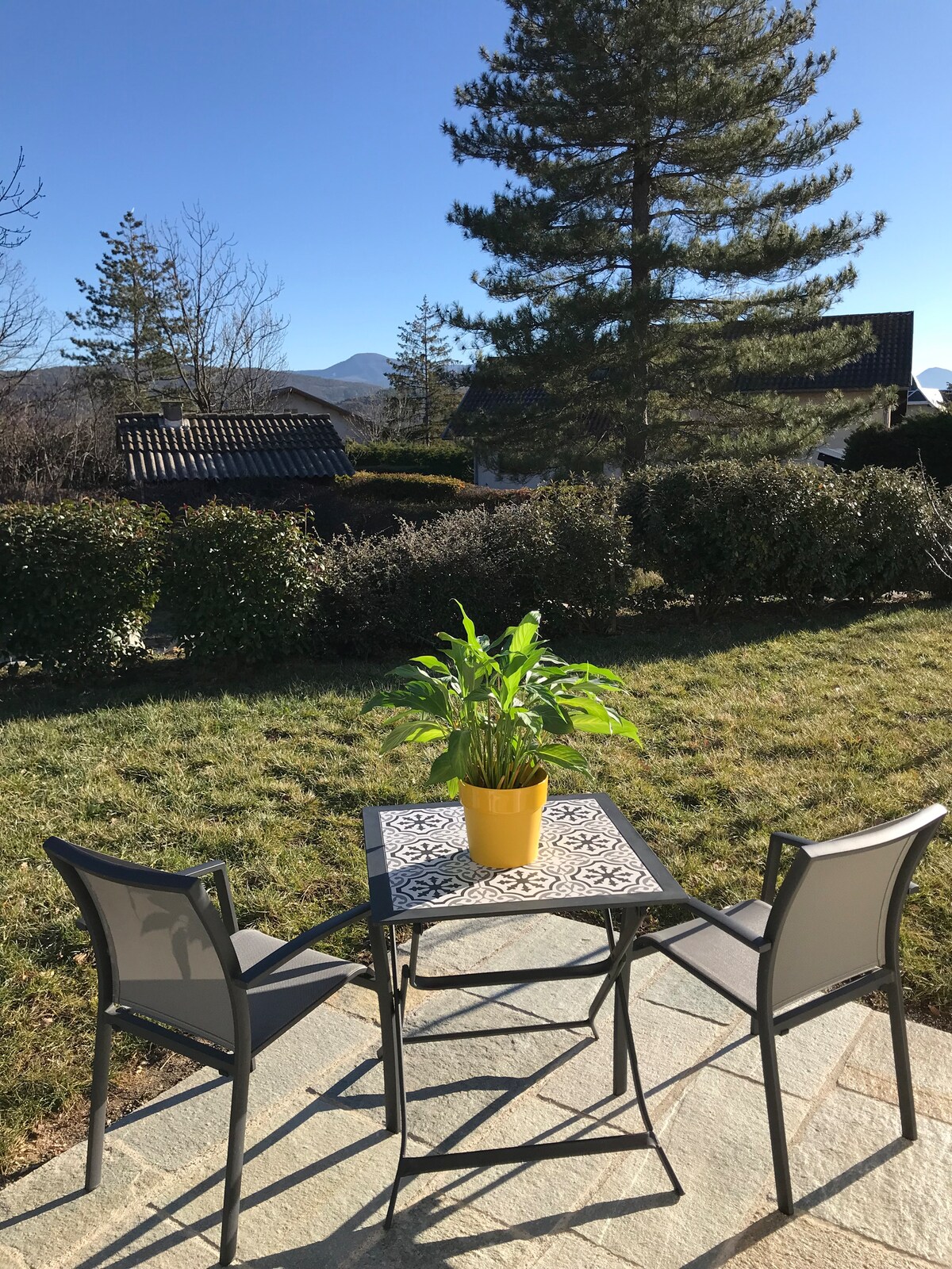 A small outdoor seating area is presented with a square table and two chairs, set on a stone surface. A potted plant adds a touch of greenery, while a backdrop of neatly trimmed hedges and distant mountains is visible under clear blue skies.