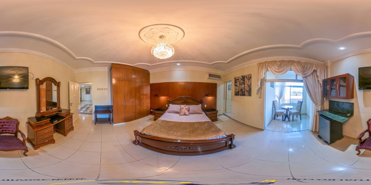 A spacious room features a comfortable bed centered under an ornate ceiling light. Wooden accents complement the decor, and a sitting area includes a small dining table beside glass doors leading to a balcony. A decorative mirror and a television are positioned against the wall.