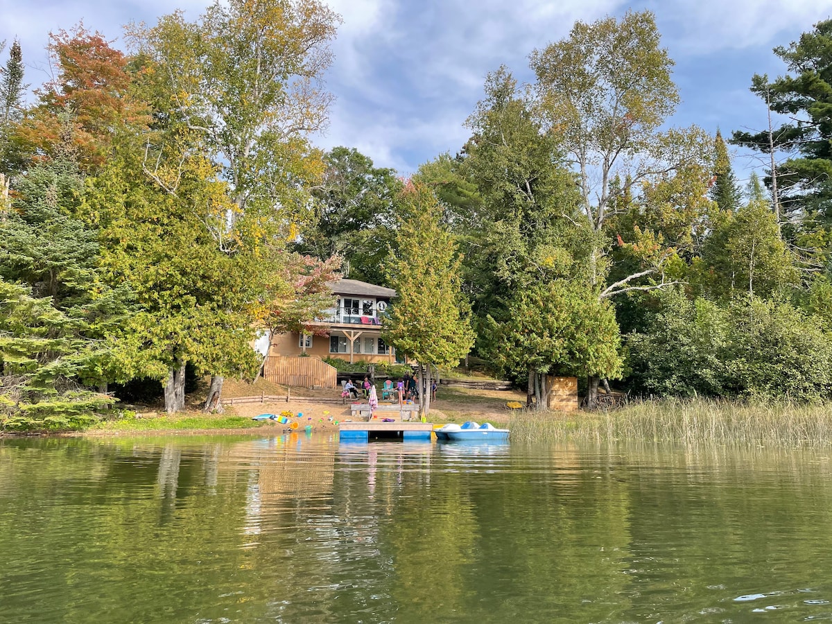 The cottage with a wooden exterior sits among lush trees by the lakefront. A dock with kayaks and paddle boats is visible, along with seating areas. The calm water reflects the greenery, creating a serene natural environment.